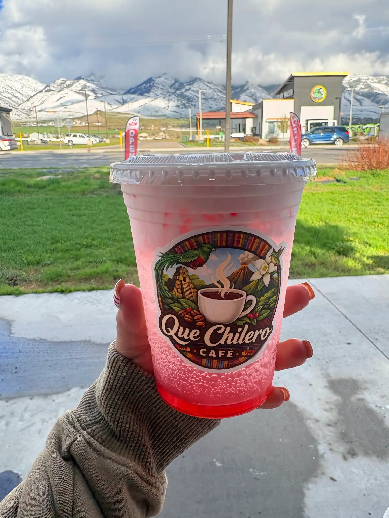 A hand holding a pink iced drink in a Que Chilero cup, snowy mountains behind